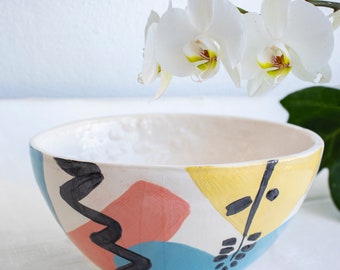 May include: A white ceramic bowl with a geometric pattern in black, yellow, pink, and blue. The bowl is sitting on a white surface with a white orchid flower hanging above it.