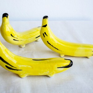 Pode incluir: Três esculturas de banana de cerâmica com detalhes pintados de preto. As bananas são amarelas e têm um acabamento brilhante.