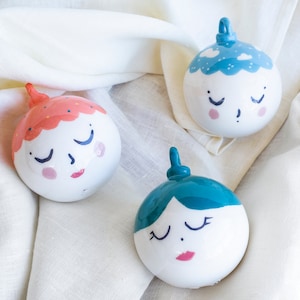 Puede incluir: Tres adornos de cerámica blanca con caras pintadas y ojos cerrados. Los adornos están decorados con diferentes colores: uno es rosa y naranja, otro es azul y otro es azul verdoso.