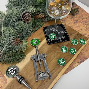 May include: A set of Dallas Stars themed wine accessories, including a bottle stopper, a corkscrew, and six wine glass charms. The charms feature the team's logo and player numbers. The background is a wooden surface with a green garland and a brick wall.