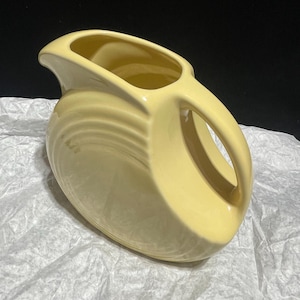 May include: A pale yellow ceramic pitcher with a curved handle and a wide spout. The pitcher has a rounded body with horizontal ridges and sits on a white surface. The pitcher is likely vintage or retro.