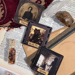 May include: Three vintage-style tarot cards with black frames and gold lettering. The cards are titled "Conjur", "Black Cat", and "Shadow". The cards are arranged on a brown surface with a book, a clear crystal, and a small bottle of amber stones.