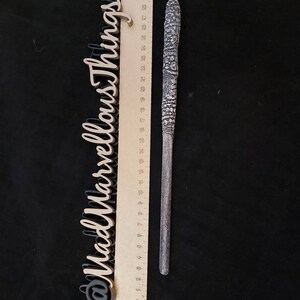 May include: A detailed, silver-coloured wand with intricate carvings and a textured handle, next to a wooden ruler. The wand is approximately 30 cm long. The background is a solid black surface. The text "@MadMarvellousThings" is also visible.