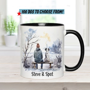 May include: White ceramic mug with a black rim and handle. The mug features a watercolor illustration of a person and a dog sitting on a bench in a snowy landscape. The text "Steve & Spot" is printed on the bottom of the mug.