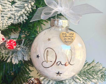 May include: A clear spherical Christmas ornament filled with white feathers, featuring the word Dad in black script and small black stars. A silver heart-shaped charm hangs from the top, inscribed with the text You are always in my heart, topped with a white sheer ribbon bow.