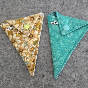 May include: Two triangular fabric pouches with floral patterns. The pouch on the left has a yellow floral pattern with a green heart charm and the text "2024" on it. The pouch on the right has a teal floral pattern with a blue button and the text "2024" on it.