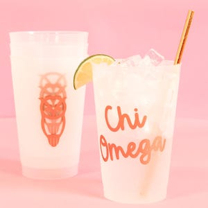 May include: Two white plastic cups with a pink background. One cup has a gold owl design and the other cup has the text "Chi Omega" in gold lettering. The cup with the text has ice and a lime wedge in it.