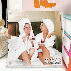 May include: Two women in white bathrobes are sitting on a bed, smiling and holding plates with cake. The photo is framed in a clear acrylic frame with a white glitter border. The letters AXQ are printed in black at the bottom of the frame.