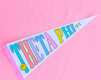 May include: A white triangular pennant with the text "THETA PHI" in colorful letters. The letters are outlined in black and have a slightly rounded font. A smiley face is in the lower left corner of the pennant.