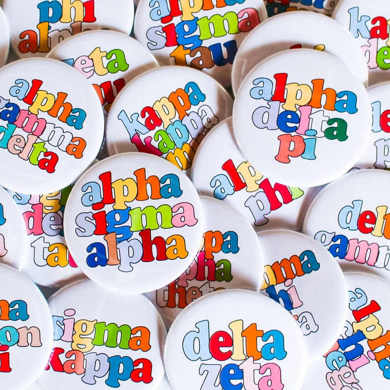 May include: A collection of colorful round buttons with Greek letters and sorority names, including Alpha Sigma Alpha, Alpha Delta Pi, Kappa Kappa Gamma, Delta Zeta, and Sigma Kappa.