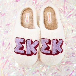 May include: White fuzzy slippers with a purple and maroon embroidered Greek letter "ΣΚ" on each slipper. The slippers have a brown label that says "DUOTTHREADS" on the inside.