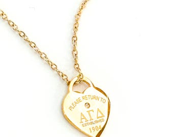 May include: Gold-colored necklace with a heart-shaped pendant. The pendant has the text "PLEASE RETURN TO" above a Greek letter symbol and the text "ESTABLISHED 1904". A small gem is set in the center of the pendant.