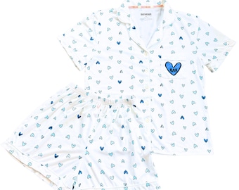 May include: White pajama set with a pattern of blue hearts. The short-sleeved top has a breast pocket with a blue heart and Greek letters. The shorts have an elastic waistband. The set is made of a lightweight fabric.