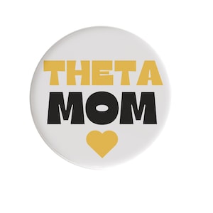 May include: A white button with the words "THETA MOM" in gold and black block letters. A gold heart is at the bottom. The button is a round shape.