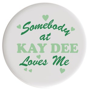 May include: White button with green text that reads "Somebody at KAY DEE Loves Me" with green hearts around the text.