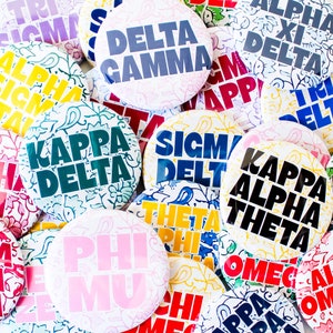 May include: A collection of colorful round buttons with various Greek letter sorority and fraternity names. The buttons feature different floral patterns and designs.