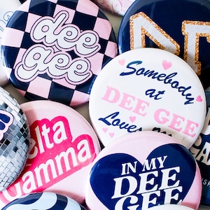 May include: A collection of colorful round pins with various designs and text. The pins feature pink, blue, white, and black colors. The text on the pins includes "dee gee", "Somebody at DEE GEE Loves", "In My DEE GEE", and "Delta Gamma".