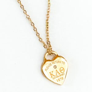 May include: Gold-toned heart-shaped pendant necklace. The pendant features the text "PLEASE RETURN TO KAO ESTABLISHED 1870" and a small gemstone. The necklace has a delicate chain.