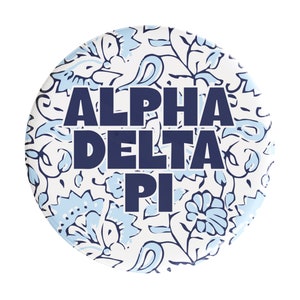 May include: A white button with a blue floral pattern and the text "ALPHA DELTA PI" in navy blue.
