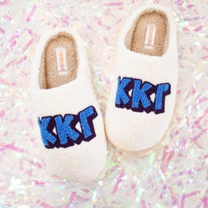 May include: White fuzzy slippers with blue and navy blue embroidered letters that spell out "KKG". The slippers have a white fluffy lining and a white label that says "DUOTHRREADS".