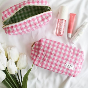 May include: Two pink and white gingham quilted makeup bags with gold zippers. The smaller bag has a green lining. The larger bag has the letters "AZ" on the front.