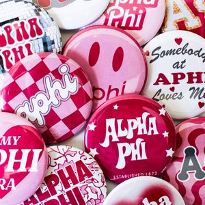 May include: A collection of red and pink Alpha Phi sorority buttons with various designs, including a checkered pattern, a smiley face, and the text "Alpha Phi" with a retro font. Some buttons also feature the text "Somebody at Alpha Phi Loves Me" and "Established 1872".