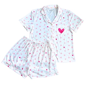 May include: White pajama set with a pink heart pattern. The set includes a short-sleeved button-up shirt with a pocket and shorts. A large pink heart is on the shirt pocket. The pajamas are made of a soft, lightweight fabric.