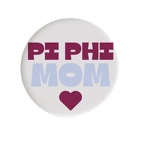 May include: A white button with the text "PI PHI MOM" in burgundy and light blue. A burgundy heart is at the bottom. The button is round and has a glossy finish.