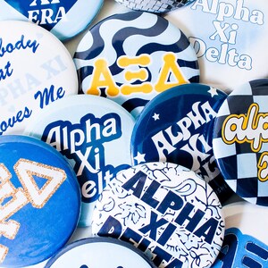 May include: A collection of blue and gold buttons with various designs, including Greek letters, stars, and checkered patterns. The buttons feature the text "Alpha Xi Delta" and "A".