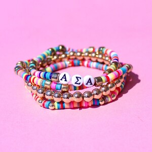 May include: Multicolored beaded bracelets with gold beads and white letter beads spelling "AΣA".