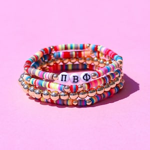 May include: Multicolored beaded bracelets with gold beads and a white bead with the letters "ΠΒΦ" in black.