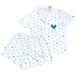 May include: White pajama set with a blue heart pattern. The short-sleeved top has a pocket with a blue heart and the initials "KKF". The shorts have an elastic waistband. The set is made of a soft, lightweight fabric.