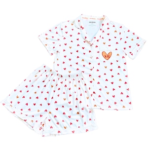 May include: White pajama set with a pattern of red and gold hearts. The short-sleeved top has a breast pocket with a heart design and the initials "K-L". The shorts have an elastic waistband. The set is made of a soft, lightweight fabric.