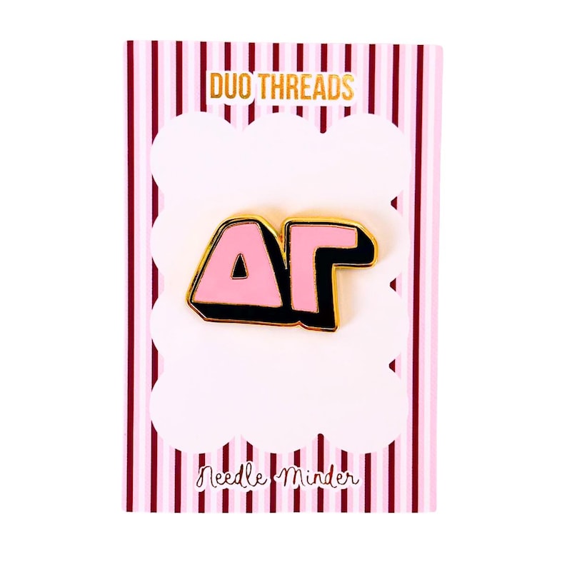 May include: A pink and black enamel pin featuring the Greek letters Delta and Gamma, mounted on a white and pink striped display card with the text DUO THREADS at the top and Needle Minder printed in black script at the bottom of the card.