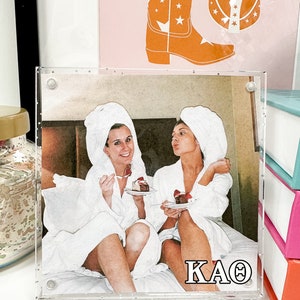 May include: A clear acrylic photo frame with a white background and a photo of two women in white bathrobes. The women are holding pieces of cake and smiling. The text "KAΘ" is printed in black at the bottom of the photo.