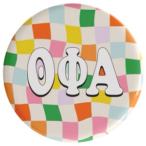 May include: A white button with a checkered pattern in shades of orange, yellow, pink, green, and blue. The letters "OΦA" are in white with a black outline and are centered on the button.