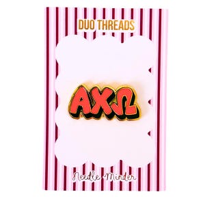 May include: A gold-toned metal enamel needle minder featuring the Greek letters Alpha Chi Omega in bright coral-red with a black outline. The pin is attached to a rectangular display card with vertical maroon stripes and the text Duo Threads Needle Minder.