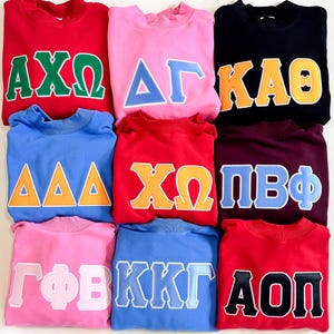 May include: Nine crewneck sweatshirts in various colors, including red, pink, blue, and black. Each sweatshirt features Greek letters in contrasting colors, such as green, orange, and blue. The sweatshirts are neatly arranged, showcasing the different color combinations.
