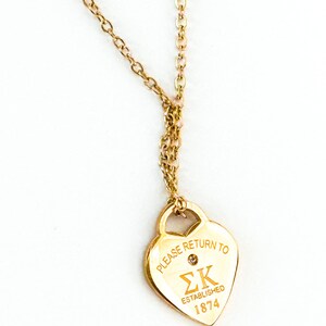 May include: Gold-colored necklace with a heart-shaped pendant. The pendant has the text "PLEASE RETURN TO ΣK ESTABLISHED 1874" and a small gem. The chain is delicate and hangs from the pendant.