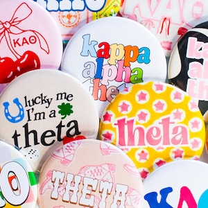 May include: A collection of colorful round buttons with various sorority names and designs. The buttons feature text such as "Kappa Alpha Theta", "Lucky Me I'm a Theta", and "Theta".