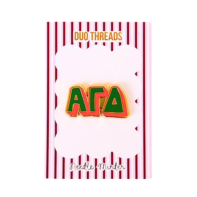 May include: A gold-toned metal needle minder featuring the Greek letters Alpha, Gamma, and Delta in green enamel with a red border. The pin is attached to a white card with pink vertical stripes, labeled DUO THREADS and Needle Minder.