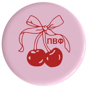 May include: A pink round button with a red cherry design. Two cherries with stems and a red bow are on a pink background. The text "PBФ" is printed in red.