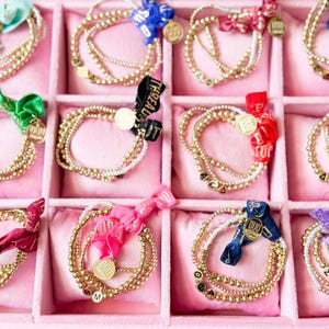 May include: A collection of gold beaded bracelets with various colored ribbons and charms, displayed in a pink jewelry box. Each bracelet features a different letter or number. The ribbons are in shades of blue, green, red, pink, and purple.