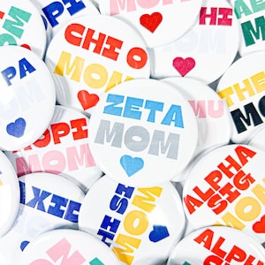 May include: A collection of white round buttons with colorful text and heart graphics. The buttons feature phrases like "ZETA MOM," "CHI O MOM," and "ALPHA SIG MOM." The text is in various colors, including blue, red, and yellow.