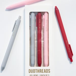 May include: A set of gel pens in a white box, with three pens inside and two pens outside. The pens are in shades of pink, red, and gray. The box reads "DUOTHREADS GEL PENS | PACK OF 3". Each pen has the text "ALPHA PHI".