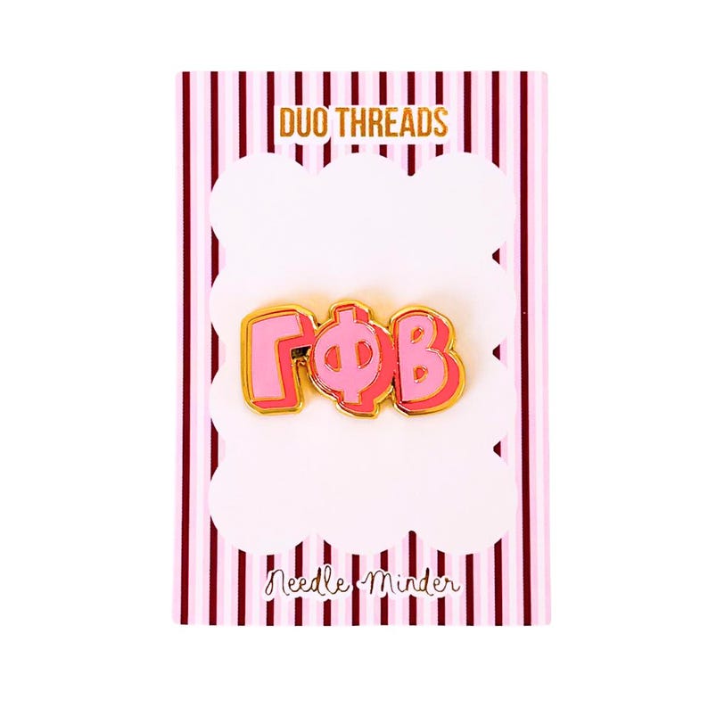 May include: A pink and gold enamel pin featuring the Greek letters Gamma Phi Beta, displayed on a white card with vertical pink stripes. The card includes the text DUO THREADS at the top and Needle Minder at the bottom in black script.