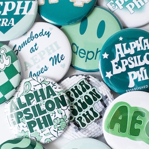May include: A collection of round buttons in shades of green and white. Some buttons feature the text "ALPHA EPSILON PHI" and "AEPHI", while others have phrases like "Somebody at AEPHI loves Me". The buttons have various designs.