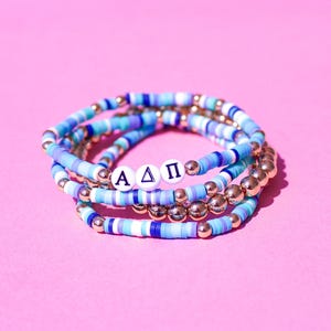 May include: Stack of four bracelets in shades of blue, white, and gold. The bracelets feature a mix of disc-shaped beads and gold-colored spherical beads. One bracelet spells out "A Δ Π" with white letter beads.