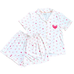 May include: White pajama set with a pink heart pattern. The short-sleeved top has a breast pocket with a large pink heart and the word "A" inside. The shorts have an elastic waistband. The set is made of a soft, lightweight material.