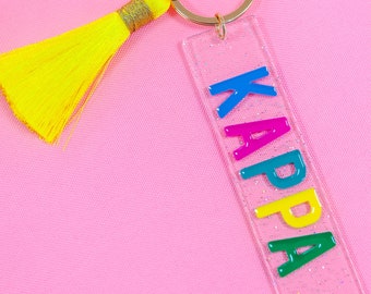 May include: A clear acrylic keychain with a colorful glitter background and the word "KAPPA" spelled out in rainbow colors. The keychain has a gold ring and a yellow tassel.